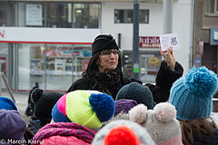 Seniorka prezentuje życzenia od uczniów. Na pierwszym planie głowy dzieci w zimowych czapkach, w tle budynki