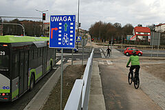 Znak "Uwaga! Rowerzysta z prawej". Na niebieskim tle informacja o konieczności ustąpienia pierwszeństwa rowerzyście. W tle skrzyżowanie z przejazdem dla rowerów oraz uczestnicy ruchu.